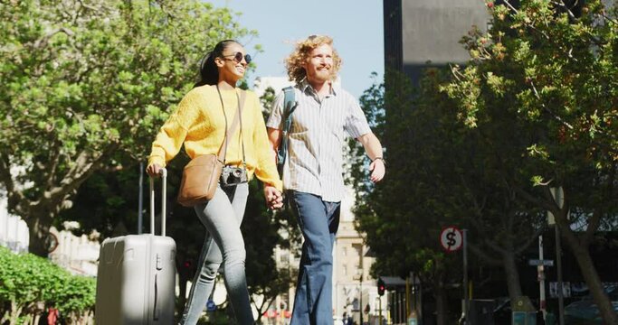 Happy diverse couple walking with luggage in sunny city street