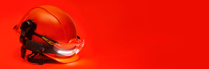 Protective goggles for eyes and construction helmet for head with earmuffs and headlamp on a red background. Construction tool and form for protection. © vadish
