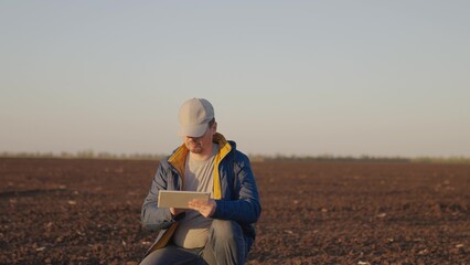 Naklejka na ściany i meble Man agronomist squats on black ground plot of plowed and cultivated field in countryside on spring day. Farmer enters data for report on soil condition into tablet. Agricultural business concept