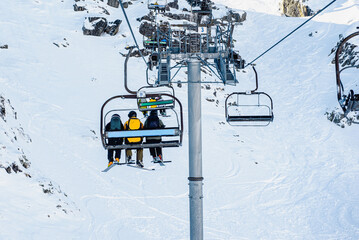 group of unrecognizable people in ski equipment, riding a chairlift up a snowy mountain on a sunny...