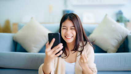 Happy young asian woman relax on comfortable couch at home texting messaging on smartphone, smiling...