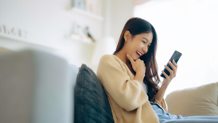 Happy young asian woman relax on comfortable couch at home texting messaging on smartphone, smiling...