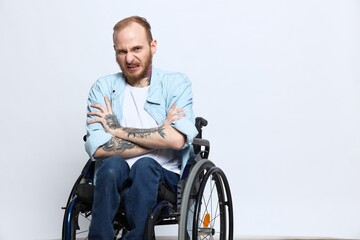 A man in a wheelchair looks at the camera anger and aggression, with tattoos on his arms sits on a gray studio background, health concept man with disabilities