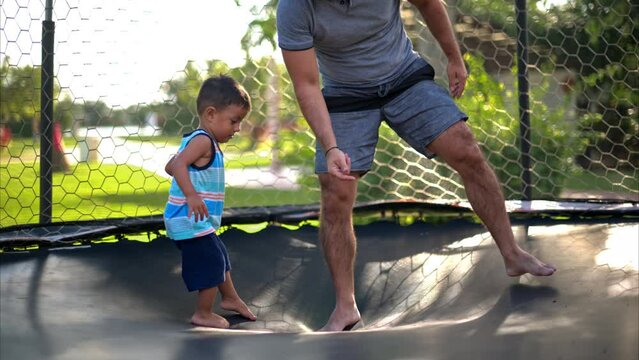 Slow motion of a young latin toddler dropping himself on a trampoline learning how to jump with his father having fun on a warm summer afternoon