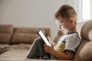 A happy little boy is sitting in a living room at home and watching cartoons on the tablet.
