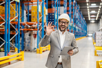 A happy warehouse manager is showing ok gesture while holding tablet and tracking inventory on it.