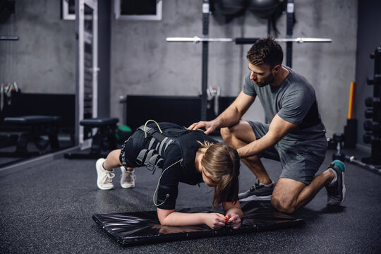 A woman in a black EMS training suit laying in a plank position on her elbows in the middle of a training center. The personal trainer corrects her body position and they prepare to start training