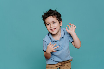 Happy curly little Italian boy in blue polo dancing, smiles against turquoise studio background....