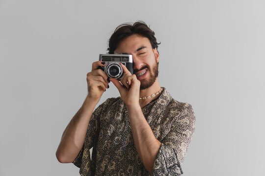 Young gay latin queer guy taking pictures with an old camera, white background