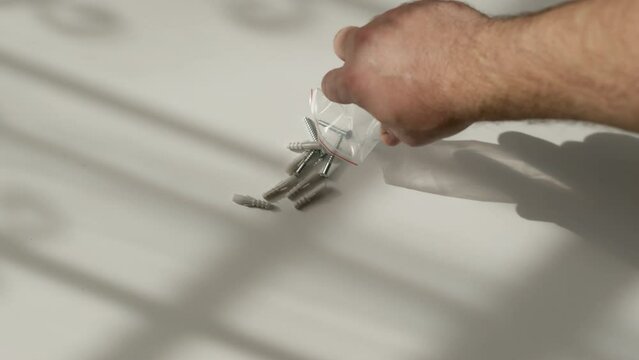 man hand pours dowels and shiny self-tapping screws onto white table surface from transparent bag. Hardware for concrete walls to hang pictures or shelves in apartment or house.