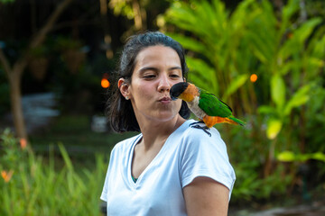 A girl kisses a green parrot that sits on her shoulder in a garden