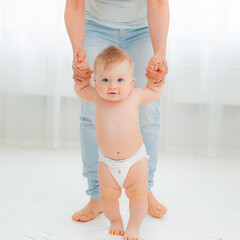 Cute little baby in a diaper learning to walk in a white room , mom holding his hands