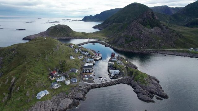 Aerial forward view of small fishing village nestled into a small sheltered cove along the mountainous coast of northern Norway on a summer day