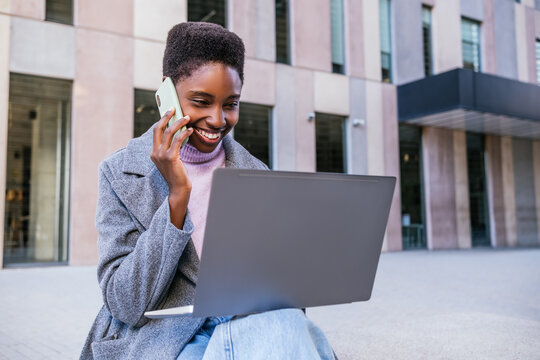 Happy African American Woman In Stylish Outerwear With Short Curly Hair Smiling And Reading Data On Laptop While Having Smartphone Conversation On City Street