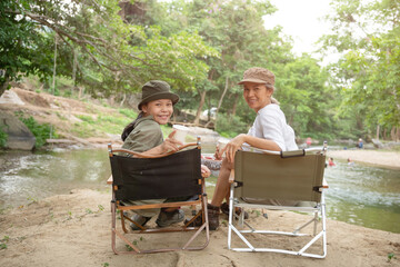Pleased happy mother and daughter sit camp chair looking out over river, sit near a camp and a tent, drink coffee in a pine forest. Camping, recreation, hiking.