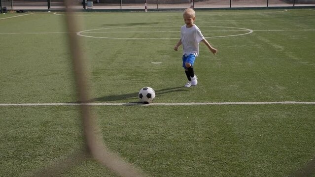 A small boy soccer player hits the goal with a soccer ball, in the foreground a blurred football net.