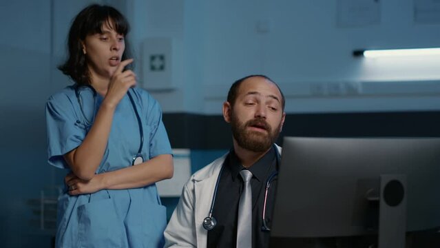 Doctor And Assistant Looking At Patient Medical Expertise On Computer Discussing Illness Symptoms Planning Medication Treatment. Clinical Staff Working After Hours In Hospital Office. Medicine Concept