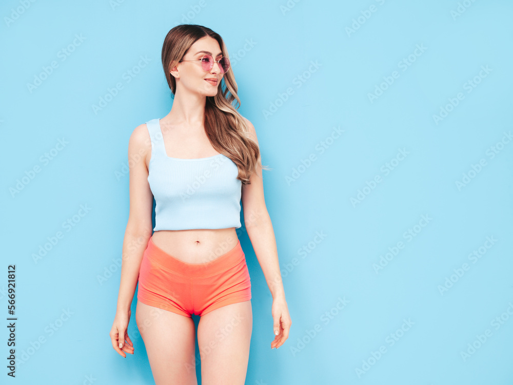 Wall mural Portrait of young beautiful smiling female in trendy summer shorts clothes. Carefree woman posing near blue wall in studio. Sexy positive model having fun indoors. Cheerful and happy. In sunglasses