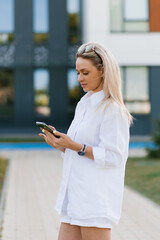 Fototapeta premium Stylish woman in a white linen suit with shorts and a shirt is shopping online on a smartphone while walking around the city in summer