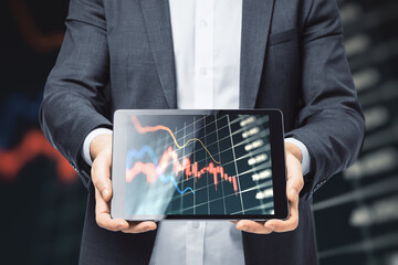 Close up of businessman hand holding tablet with glowing falling forex chart on blurry dark grid...