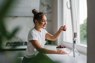 Young woman nutritionist blogger conducts a consultation or an open lesson by video link telling about the benefits of fresh berries, sitting in the kitchen with a plate of cherries