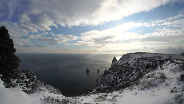 Snow covered rocky cliffs over sea. Winter landscape over the sea bay. Juniper tree under snow. Concept of winter holidays, travel and recreation. Cape Fiolent in Balaklava, Sevastopol, Crimea