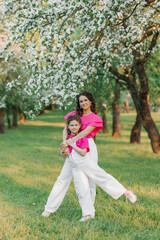 Naklejka na ściany i meble A sweet young woman in white trousers and a bright pink blouse with her daughter is walking through a spring park or garden among the flowering trees of an apple tree and hugs her