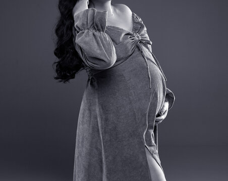 A pregnant woman in a dress. A pregnant woman in a gray dress stands on a gray background with a place for text. Studio shooting of pregnancy. Black and white photo. Black and white image of a person.
