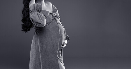 A pregnant woman in a dress holds her belly. A pregnant woman with long hair stands on a gray background with a place for text. Pregnancy. Pregnancy studio shooting.