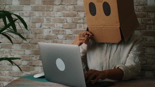 Catfish online crime concept with person and cardboard to hidden identity face in front of a laptop computer working and thinking. Unhappy worker writing on notebook indoor. Modern lifestyle alone