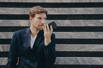 Male executive worker dressed formally holds smartphone near mouth talks on speakerphone