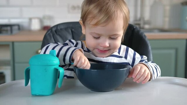 One year old hungry baby girl in striped casual clothes sits at white table in a gray highchair and eats herself with spoon. Blurred dining room background. Healthy eating for kids. Child's nutrition.