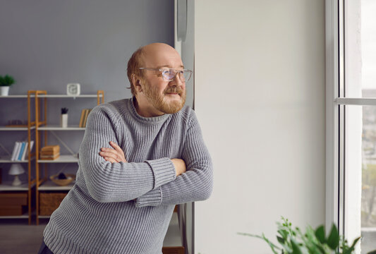 Happy Retired Senior Man Looking Out The Window. Portrait Of A Bald Bearded Old Man In Glasses Standing With His Arms Crossed By The Window At Home, Looking Out And Thinking