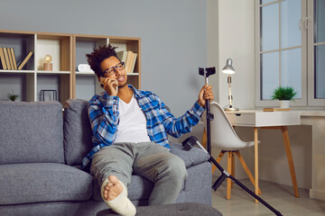 Handsome young man with fractured leg sitting on sofa talking on cell phone. Cheerful casually...