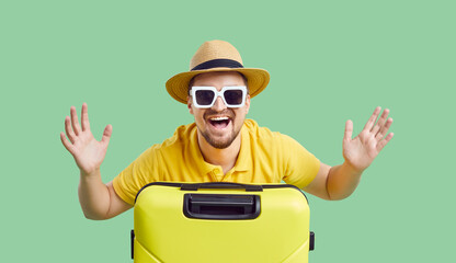 Portrait of overjoyed young man traveler in straw hat and sunglasses with suitcase excited about vacation. Smiling male tourist with baggage ready for summer holidays. Green studio background.