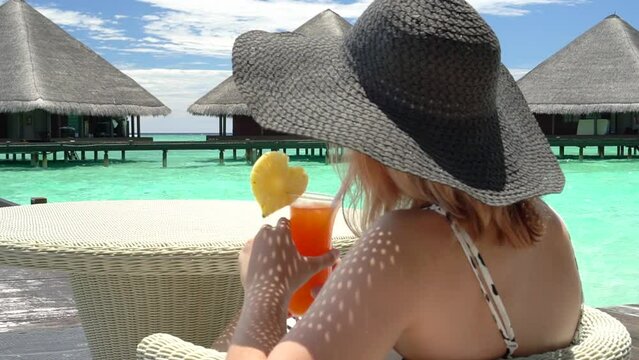 A young happy girl in a big black beach hat sits at a table and holds a bright yellow ceta cocktail with a pineapple heart in her hands against the backdrop of water villas in the Maldives. Close-up