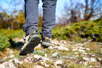 Legs of a hiker in trekking boots walking in the mountains closeup shot. Feet of walking tourist wearing trekking shoes on a rocky road captured from behind. Hiking male wearing pants and boots walk