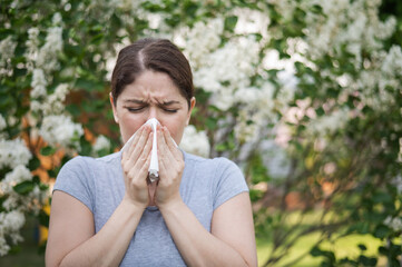 Caucasian woman suffers from allergies and blows her nose into a napkin while walking in the park.  - obrazy, fototapety, plakaty