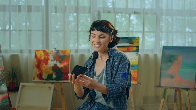 In the art studio beautiful and charismatic artist woman taking some selfies pictures with her smartphone while sitting on the chair