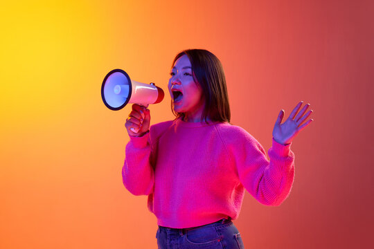 News, information. Young girl in pink sweater talking in megaphone over gradient orange background in neon light. Concept of emotions, facial expression, youth, lifestyle, inspiration, sales, ad