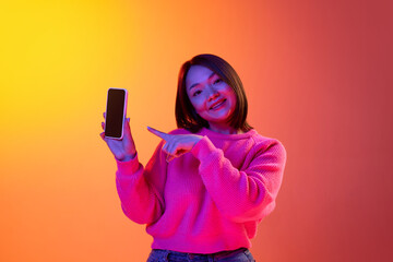 Online shopping. Young girl in pink sweater smiling, pointing on phone screen over gradient orange...