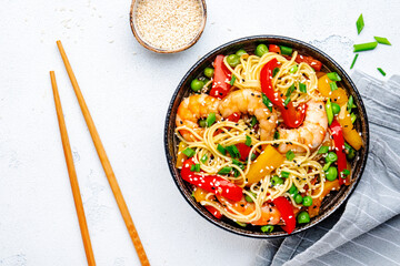 Stir fry noodles with shrimps, red and yellow paprika, green pea, chives and sesame seeds in ceramic bowl. Asian cuisine dish. White stone kitchen table background, top view