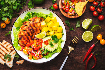 Grilled chicken salad with avocado, mango, tomato salsa, cilantro and lettuce in mexican style, rustic wooden table background, top view