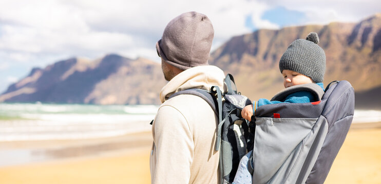 Young father carrying his infant baby boy sun in backpack on windy sandy beach of Famara, Lanzarote island, Spain. Family travel and winter vacation concept. Parenting, fatherhood, father's day
