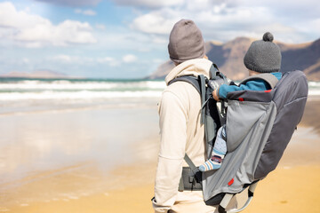 Young father carrying his infant baby boy sun in backpack on windy sandy beach of Famara, Lanzarote island, Spain. Family travel and winter vacation concept. Parenting, fatherhood, father's day