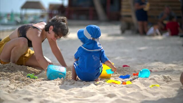 Cute little boy playing on the sand with beach toys and his auntie in a sunny day