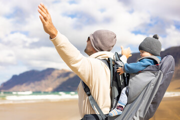 Young father rising hands to the sky while enjoying pure nature carrying his infant baby boy sun in backpack on windy sandy beach of Famara, Lanzarote island, Spain. Family travel concept