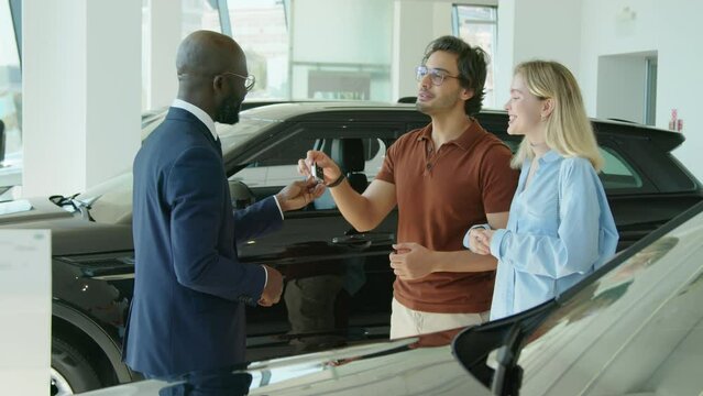 African American salesperson working in dealership center passing new car key to happy young couple