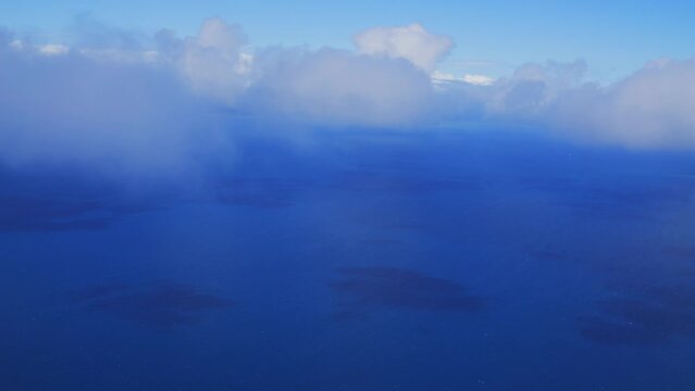 A view from a plane captures the breathtaking sight of going through the clouds above the sea, showcasing the beauty of the white fluffy clouds contrasting with the deep blue of the ocean.