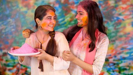 Obraz na płótnie Canvas Indian sister celebrating Holi by applying color on cheeks - Tradition smiling faces pink color. Video of the two attractive sisters painted in the colors of Holi - Sisterhood happiness relatio...